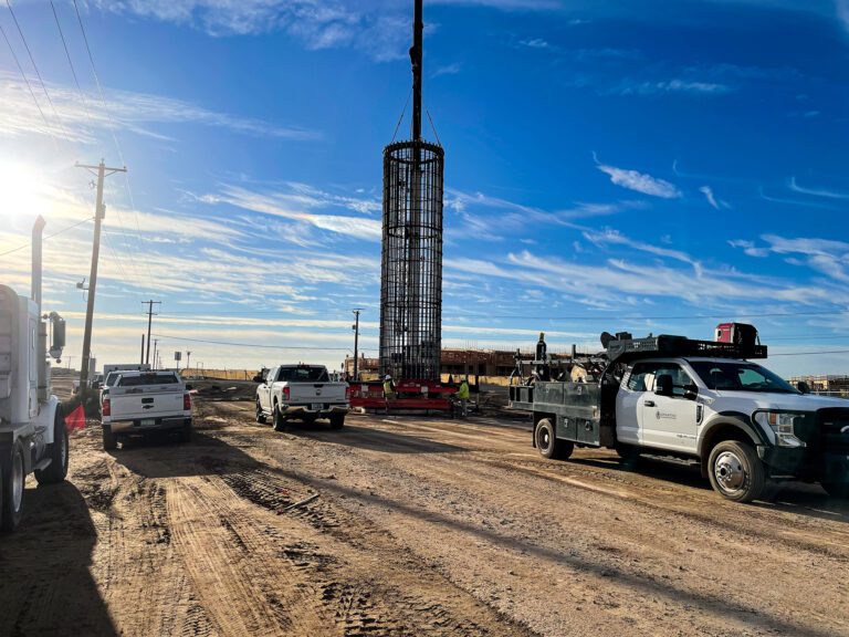 Construction site with vehicles and a tall reinforced concrete column being lifted by a crane under a clear blue sky, near the installation of an Able Pfister Ball Transmission Line.