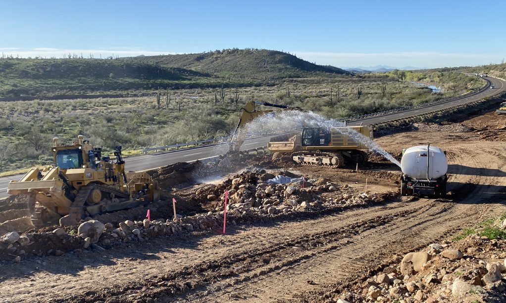 Bulldozer at a road construction site moving earth while a water truck sprays to control dust, showcasing Civil Engineering News and Events.