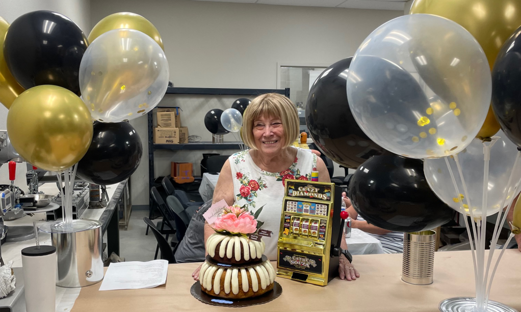 A smiling Cathy Souza celebrates her retirement with a cake, flowers, balloons, and an award in a decorated room.